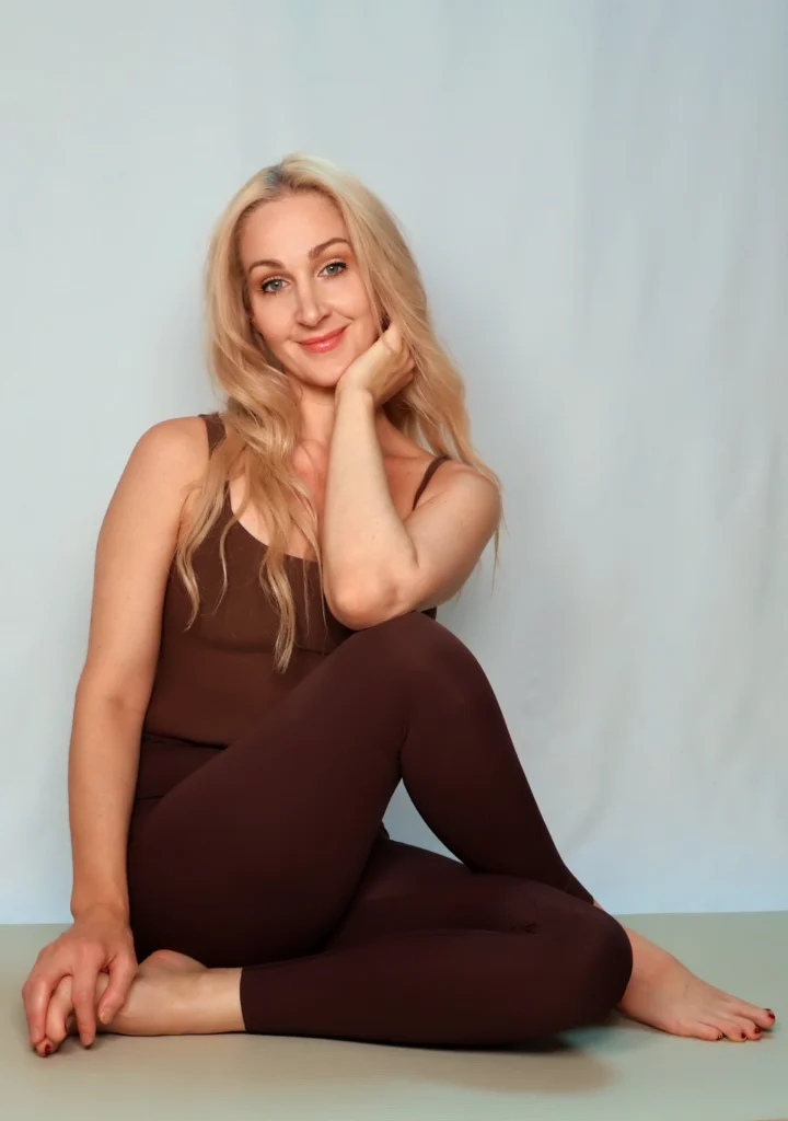 Ly Tolberg Joogaõpetaja portreefoto. Pikemate blondide juustega naeratav naine istub matil mahedas pruunis joogakostüümis ja toetab kätt pehmelt põsele, taustaks on helge ja minimalistlik hele sein.